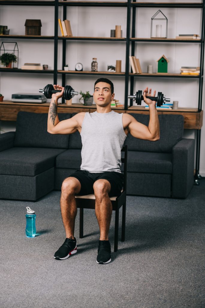 tattooed mixed race man exercising with dumbbells on chair in home gym